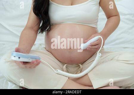 primo piano di una giovane donna incinta che utilizza il dispositivo droppler fetale per ascoltare il battito cardiaco del bambino Foto Stock