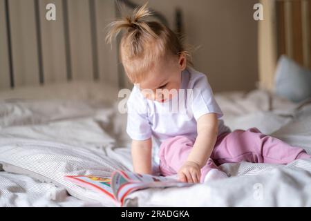 Bambina che legge, guarda le foto nel libro a letto a casa, un bambino curioso vuole leggere una nuova fiaba. Foto Stock