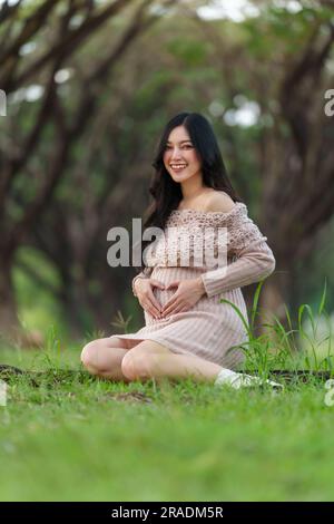 donna incinta felice seduta nel parco e mostra la forma del cuore sulla pancia Foto Stock