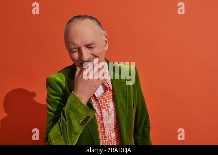 felice invecchiamento, felicissimo uomo anziano che copre la bocca con la mano e ride con gli occhi chiusi su sfondo rosso arancione, blazer verde velluto, camicia alla moda, Foto Stock