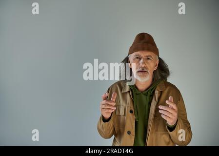 turbato e preoccupato, uomo barbato anziano con berretto e giacca marrone che gestiva e guardava la macchina fotografica su sfondo grigio, vestiti hipster, gente anziana Foto Stock