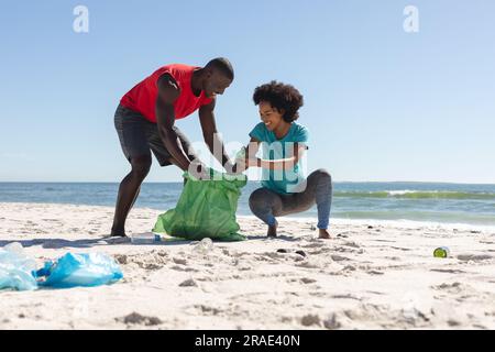 Felice coppia afro-americana che pulisce la spiaggia soleggiata e raccoglie i rifiuti Foto Stock