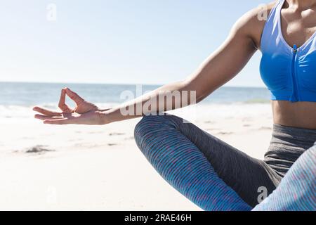 Sezione intermedia di una donna afroamericana in forma che pratica meditazione yoga seduto sulla spiaggia soleggiata Foto Stock