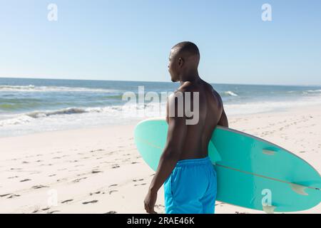 Un uomo afro-americano che porta la tavola da surf sulla spiaggia soleggiata, affacciato sul mare Foto Stock