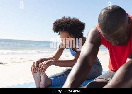 Allenati con una coppia afroamericana che pratica yoga, seduto e distendendosi sulla spiaggia soleggiata Foto Stock