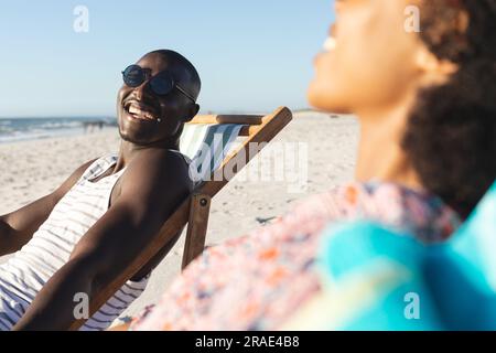 Felice coppia afro-americana in occhiali da sole seduti su sdraio ridendo sulla spiaggia soleggiata Foto Stock