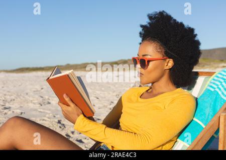 Felice donna afro-americana con occhiali da sole seduta su sdraio a leggere un libro sulla spiaggia soleggiata Foto Stock