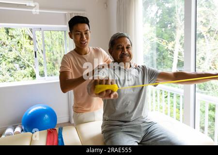 Fisioterapista maschile felice eterogeneo che consiglia e paziente di sesso maschile anziano che utilizza la fascia da ginnastica Foto Stock