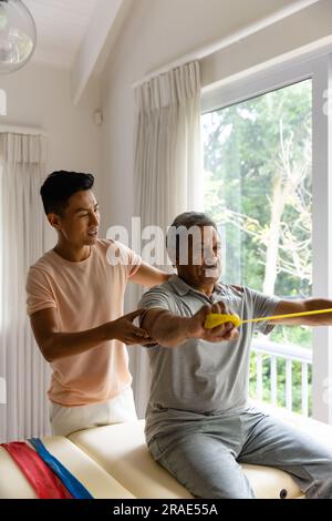 Fisioterapista maschile felice eterogeneo che consiglia e paziente di sesso maschile anziano che utilizza la fascia da ginnastica Foto Stock