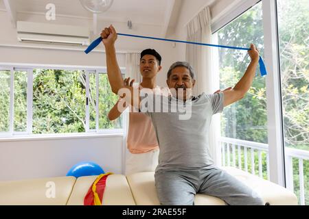 Fisioterapista maschile felice eterogeneo che consiglia e paziente di sesso maschile anziano che utilizza la fascia da ginnastica Foto Stock