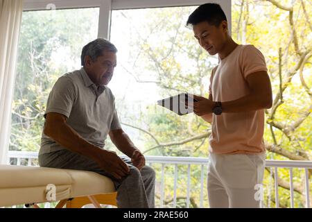 Fisioterapista maschile eterogeneo felice che esamina pazienti di sesso maschile anziani utilizzando la tavoletta Foto Stock