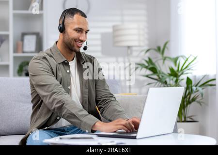 Giovane studente ispanico seduto a casa su un divano che indossa cuffie e studia online su un computer portatile. Foto Stock
