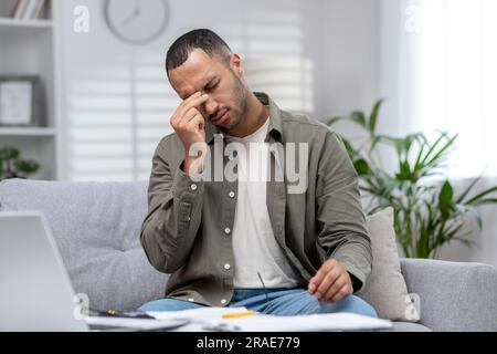 Giovane studente maschio ispanico che studia a distanza a casa. Si siede stancamente sul divano di fronte al portatile, tiene gli occhiali, strofina gli occhi, si riposa. Foto Stock