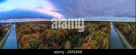 Immergiti nel vivace arazzo autunnale mentre la fotocamera cattura un'incantevole vista aerea di un fiume o di un canale che scorre attraverso i boschi, adornati da colorate foglie autunnali, il tutto contro un suggestivo cielo nuvoloso. La tavolozza della natura prende vita mentre le foglie si trasformano in un caleidoscopio di tonalità calde, dipingendo il paesaggio con sfumature di rosso, arancione e oro. Il tranquillo corso d'acqua si snoda attraverso i pittoreschi boschi, riflettendo i colori accattivanti sopra. Il suggestivo cielo nuvoloso aggiunge profondità e contrasto, creando uno sfondo suggestivo che esalta la bellezza dell'autunno Foto Stock