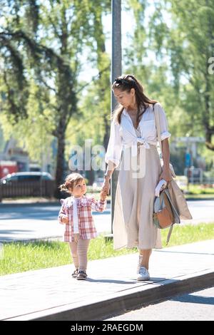 Madre e figlia piccola che si tengono per mano mentre camminano insieme su un marciapiede. Giorno estivo di sole. Shot verticale. Foto Stock