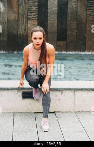 Ritratto all'aperto di una giovane bella donna in forma che si allunga sulle gambe, indossa abbigliamento sportivo alla moda, modello di atleta in posa sullo sfondo urbano, moda sportiva Foto Stock