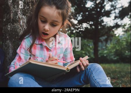 Ritratto da vicino Caucasica bella bambina, prima elementare, studentessa della scuola elementare, seduta sotto l'albero e leggendo un libro, facendo i compiti a casa Foto Stock