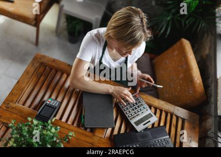 Giovane imprenditrice seduta alla scrivania e digitando al computer mentre chiacchiera con lo smartphone. Felice fiorista femminile in verde floreale centratura su l Foto Stock