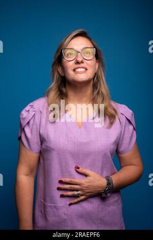 Bella donna brasiliana indossando occhiali indossando uniforme nutrice lilla con mano sul ventre guardando in alto. persona positiva. Isoald su sfondo blu. Foto Stock