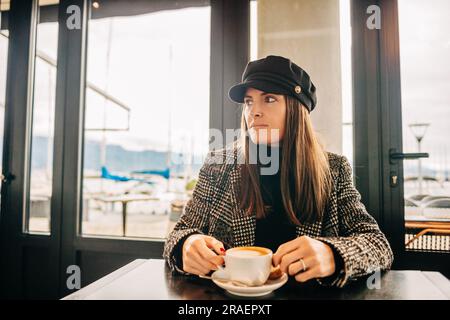 Giovane bella donna che beve un caffè in un bar, indossa una giacca in tweed e un cappello da ragazzo baker con cordoncino nero Foto Stock