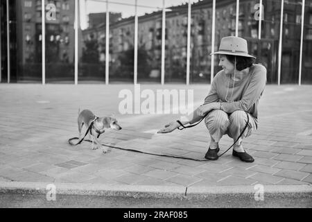 Giovane adulta in città con un cane seduto e abbracciato, sorridente. Accarezzare il cane, nutrirsi, divertirsi Foto di alta qualità Foto Stock