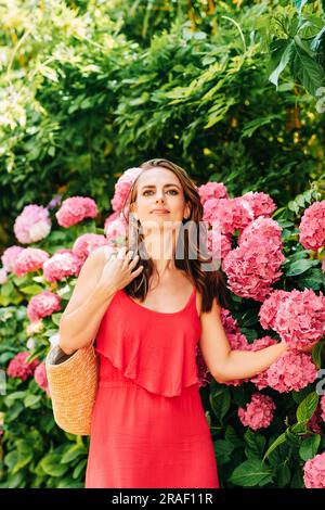 Ritratto all'aperto di una bella donna in posa con fiori di ortensia rosa, indossando un vestito rosso Foto Stock