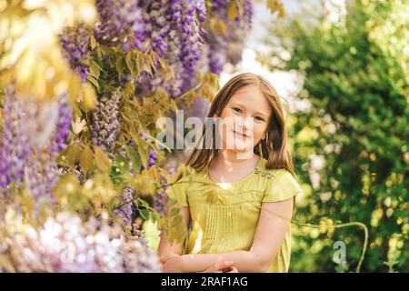 Elegante ritratto all'aperto di una graziosa bambina di 8-9 anni, accanto a splendidi fiori di glicine viola, che indossa una camicetta verde Foto Stock