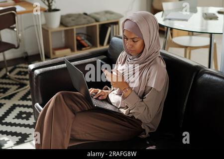 Giovane operaia musulmana con un computer portatile in ginocchio seduta su un comodo divano in salotto e scorrimento nel telefono cellulare Foto Stock