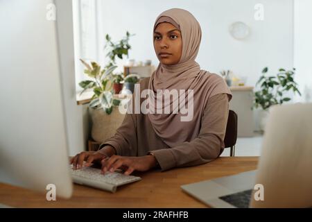 Giovane programmatore donna seria che guarda lo schermo del computer mentre analizza o decodifica dati o sviluppa nuovo software sul posto di lavoro Foto Stock