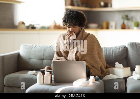 Un uomo indiano malato coperto di coperta che guarda il laptop e starnutisce il naso Foto Stock