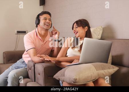 Indiano coppia felice mangiare insalata e dieta sana seduti su un divano tenendo un portatile navigare online. Ascolto di musica Foto Stock