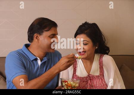 Felice indiano salute conscio marito moglie seduto insieme avendo una ciotola di insalata verde seduto sul divano in un soggiorno Foto Stock