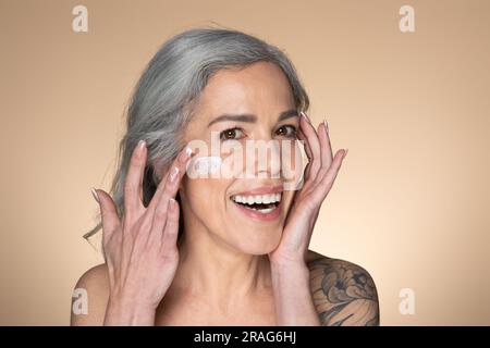 Ritratto di una donna anziana dai capelli grigi con crema idratante sulle guance, che prova un nuovo prodotto di bellezza Foto Stock