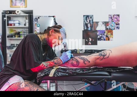 Un bellissimo maestro creativo con i guanti sta lavorando per un nuovo tatuaggio a gambe grandi per un cliente. Foto di alta qualità Foto Stock