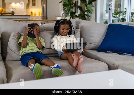Fratello e sorella afro-americani che usano tablet e smartphone mentre sono seduti sul divano nel soggiorno Foto Stock