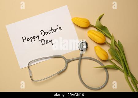Scheda con frase Happy Doctor's Day, stetoscopio e tulipani gialli su sfondo beige, piatto Foto Stock