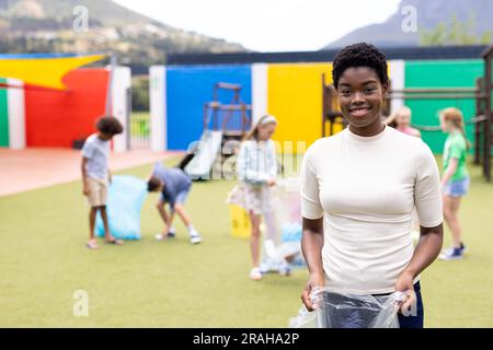 Ritratto della sorridente maestra afroamericana che raccoglie riciclaggio nel cortile della scuola, spazio per le copie. Istruzione, inclusività, scuola elementare e lear Foto Stock