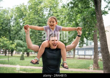 Una bambina che fa piggybacks sul padre, cavalca sulla spalla del padre nel parco. Tempo di Unione familiare felice, concetto di paternità Foto Stock