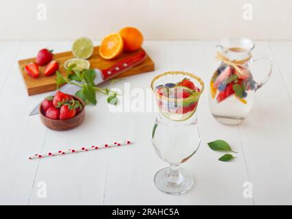 Una colorata composizione di arance fresche, fragole, mirtilli, lime e menta, servita su un tavolo di legno bianco, con vetro alto e una caraffa Foto Stock