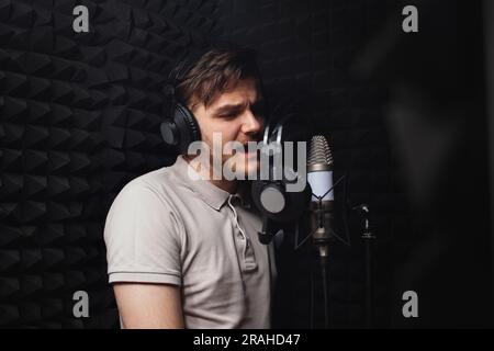 l'uomo canta nel microfono, nello studio di registrazione professionale, nelle cuffie. Pareti nere morbide, isolamento acustico. cantante registra la sua canzone. Foto Stock