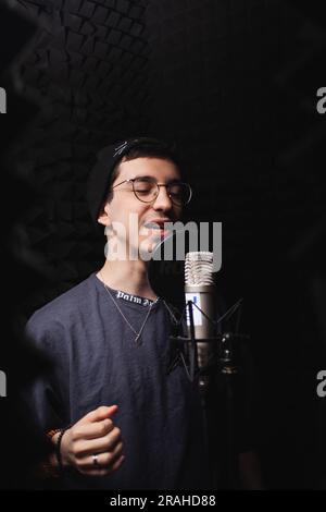 l'uomo canta nel microfono, nello studio di registrazione professionale, nelle cuffie. Pareti nere morbide, isolamento acustico. cantante registra la sua canzone. uomo con gli occhiali Foto Stock