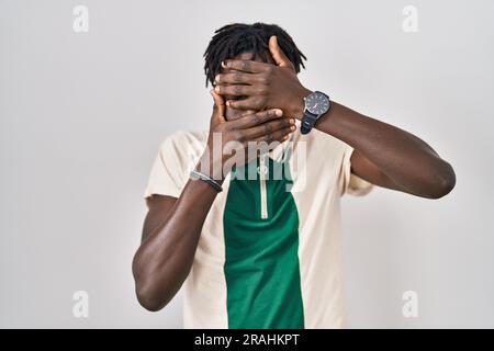 Uomo africano con dreadlocks in piedi su uno sfondo isolato che copre occhi e bocca con le mani, sorpreso e scioccato. nascondere le emozioni Foto Stock