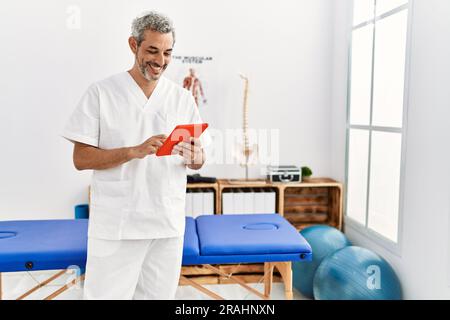 Fisioterapista uomo dai capelli grigi di mezza età sorridente sicuro usando il touchpad alla clinica di riabilitazione Foto Stock