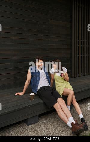 Un'allegra bruna che indossa un elegante outfit estivo con caffè per andare a parlare con il fidanzato mentre si siede vicino a una casa di legno Foto Stock