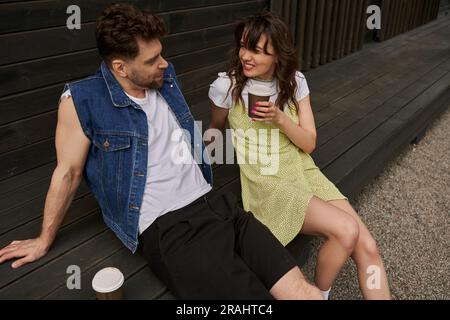 Uomo barbato sorridente ed elegante in un gilet in denim che parla con una bruna fidanzata in abito da sole che tiene il caffè per andare a sedersi vicino a una casa di legno in campagna Foto Stock