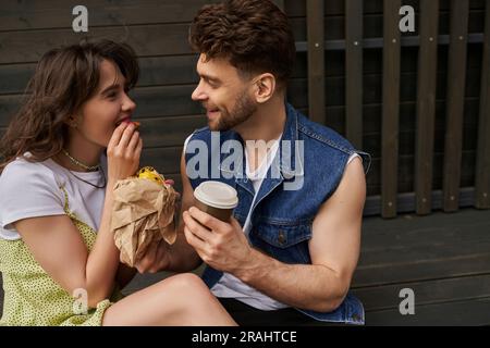 Uomo barbato sorridente in un gilet in denim che tiene il caffè per andare a parlare con la fidanzata in un vestito estivo mangiando panino fresco e seduto vicino alla casa di legno a ba Foto Stock