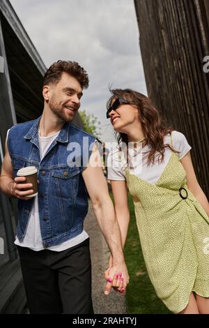 Allegra coppia romantica con eleganti abiti estivi che si tengono per mano e caffè da andare mentre si parla e si cammina tra le case di legno in un ambiente rurale Foto Stock