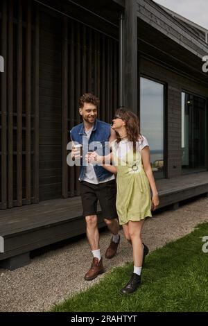 Coppia alla moda ed elegante in estate abiti e stivali che tengono in mano il caffè da asporto e parlano mentre camminano insieme vicino a una casa di legno in un ambiente rurale, o Foto Stock