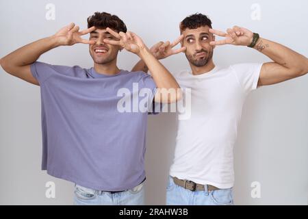 Coppia omosessuale gay in piedi su sfondo bianco facendo simbolo di pace con le dita sul viso, sorridendo allegro mostrando la vittoria Foto Stock