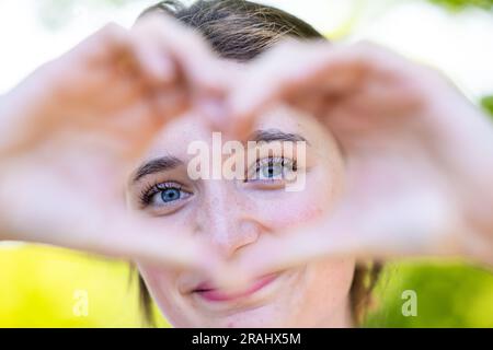 Ammira la bellezza accattivante e la gioia radiosa mentre una giovane donna crea un simbolo del cuore con le mani, osservandolo con un aspetto di puro incanto. Questo ritratto la cattura in un momento di sincera connessione, mentre l'amore e la gioia risuonano dentro di lei. Il simbolo del cuore rappresenta il suo spirito aperto e compassionevole, simboleggiando le profondità delle sue emozioni. Il suo sguardo accattivante riflette la bellezza genuina che deriva dall'abbracciare amore e gioia. Osserva il potere della sua espressione mentre crea una cornice visiva di affetto, invitandoti a sentire il calore e la positività che irradia da lei. Hea Foto Stock